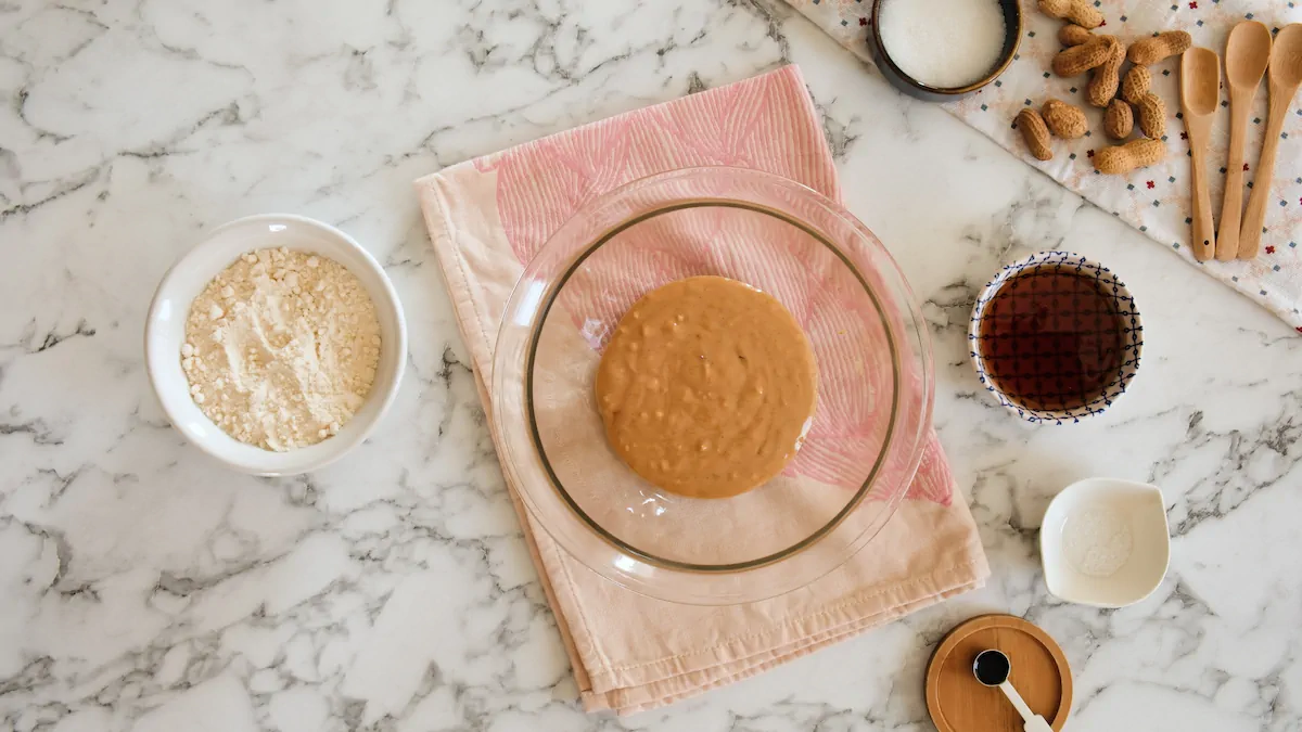 A mixing bowl with peanut butter in the center surrounded by sugar-free maple syrup, vanilla extract, coconut flour and sea salt flakes in different bowls..