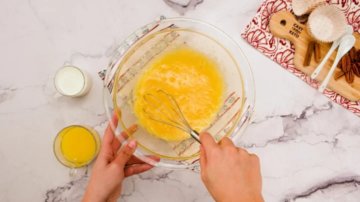 Whisking the eggs and granulated erythritol in a large bowl alongside melted butter and almond milk in different glasses.