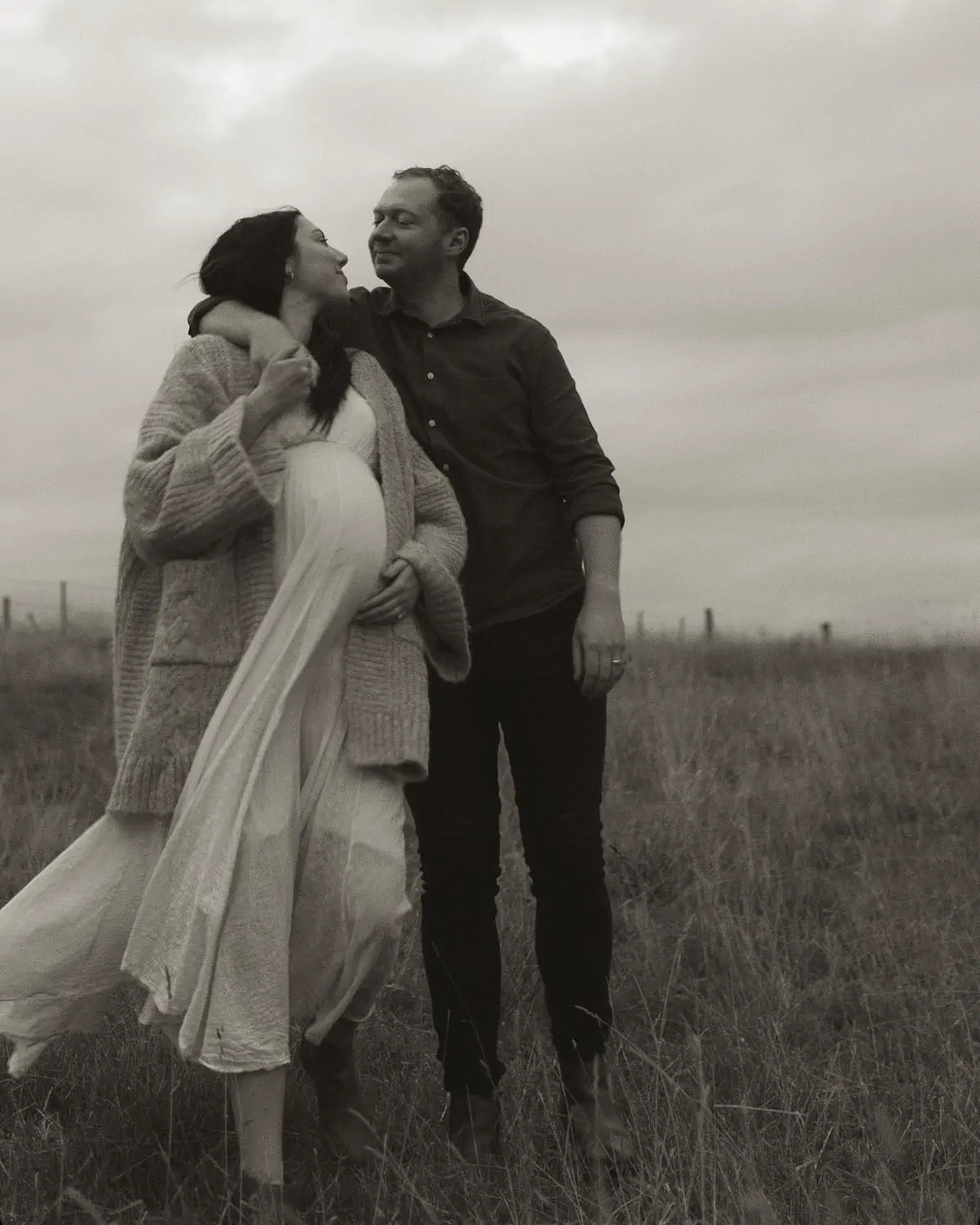 A black and white picture of Frank with his pregnant wife Georgie in the fields, his hand over her shoulder and looking into each other's eyes.