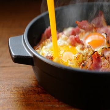 A cast iron pan with eggs , bacon and steam coming out of it.