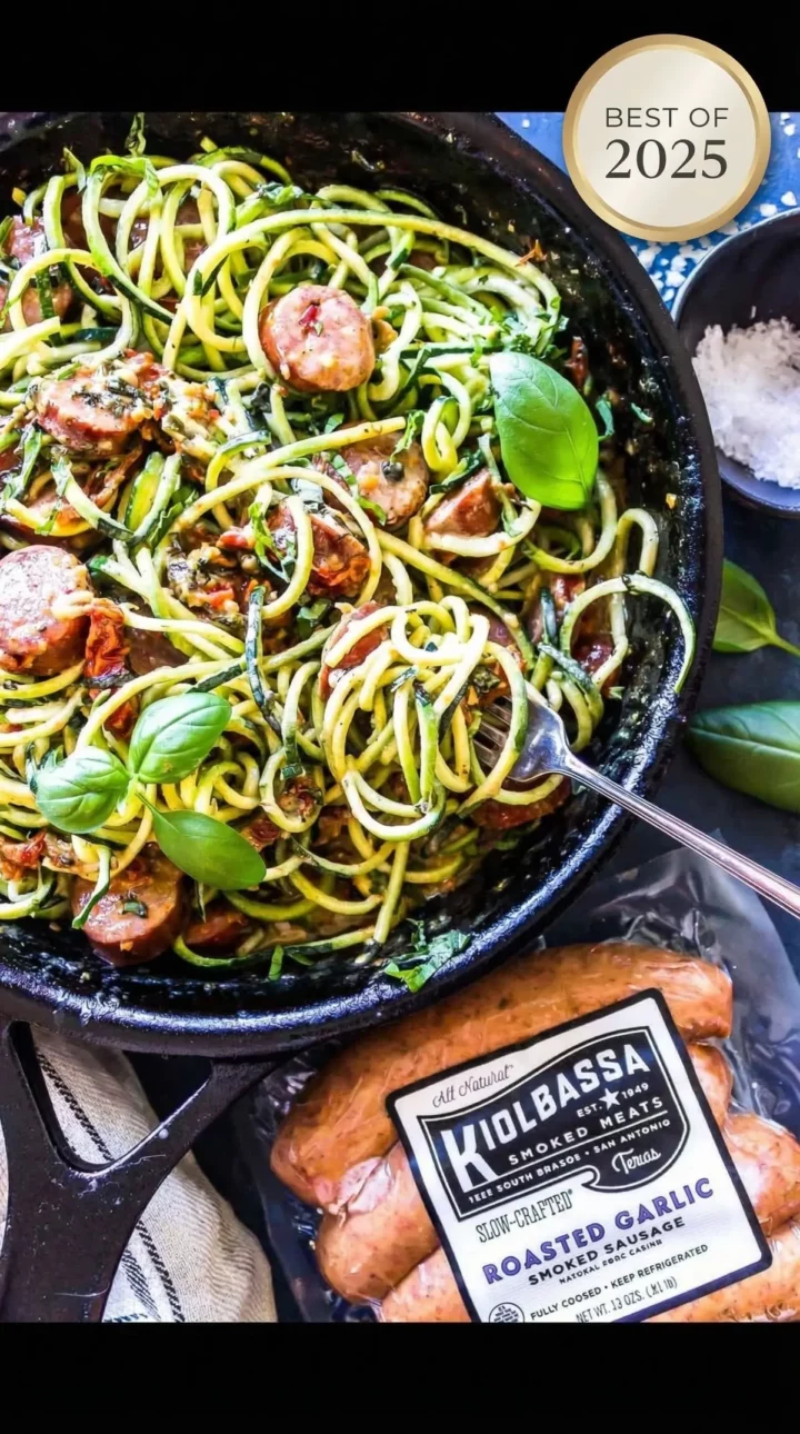 Skillet of spiralized zucchini noodles tossed with sausage slices, herbs, and garlic, with a package of smoked sausage in the foreground