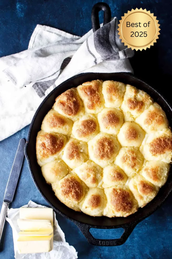 Golden brown dinner rolls baked in a cast iron skillet, served with sliced butter and a butter knife on a dark blue background.
