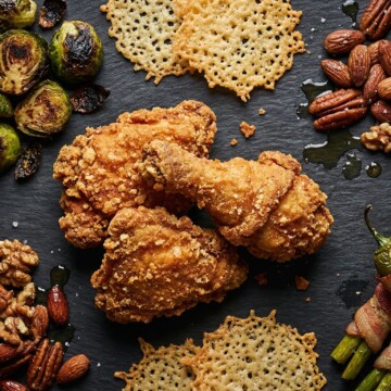 Assorted crispy and texture-rich keto foods like fried chicken, roasted Brussels sprouts, cheese crisps, and mixed nuts on a dark background