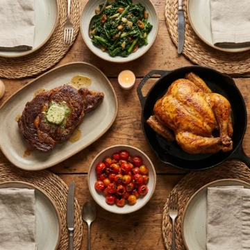 A top-down view of a rustic dinner table featuring steak, salmon, chicken, and vegetable sides
